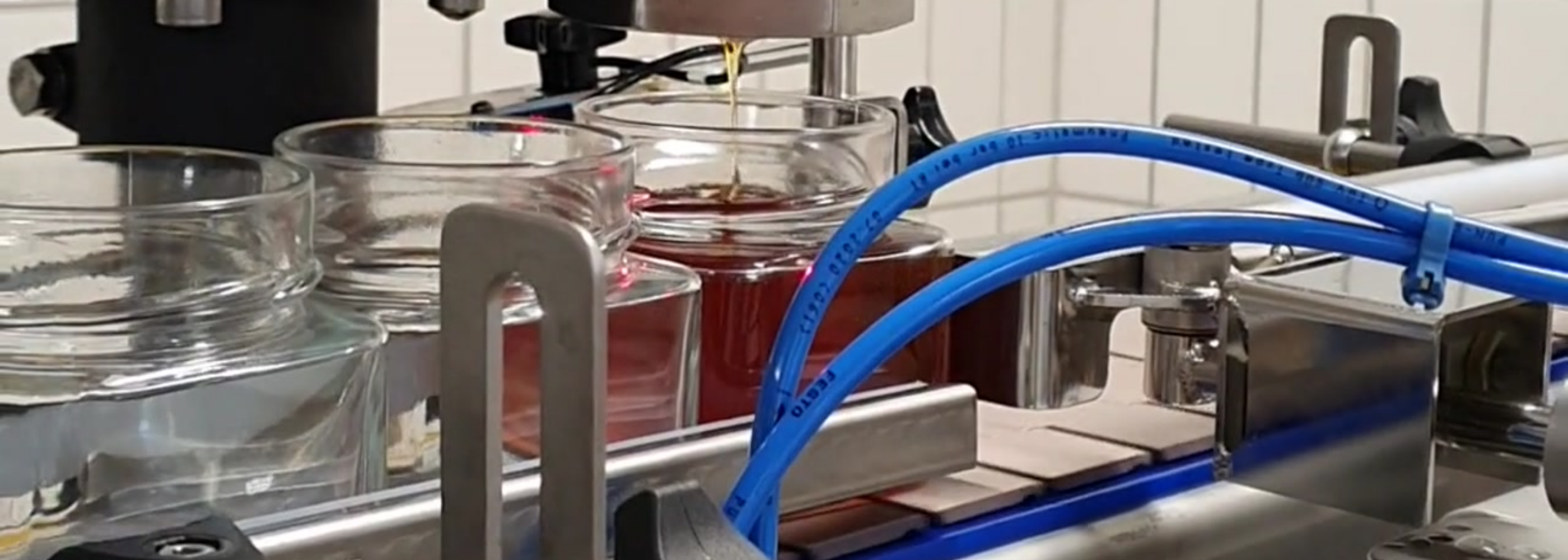 Eine Nahaufnahme einer automatisierten Abfüllanlage in einer Fabrik. Mehrere Honiggläser (oder Sirupgläser) bewegen sich auf einem Förderband. Einige Gläser sind bereits mit bernsteinfarbenem Honig/Sirup gefüllt, während andere noch leer sind. Im Vordergrund dominiert ein blaues Pneumatikrohr die Szene. Das Bild symbolisiert Lebensmittelproduktion und Abfüllung.